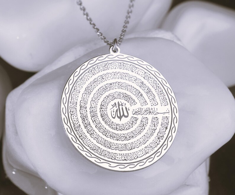 Pu&ograve; includere: Un ciondolo circolare color argento con un'intricata calligrafia araba incisa a spirale. Il centro mostra la parola Allah, circondata da linee di testo concentriche, rifinito con un bordo intrecciato decorativo.