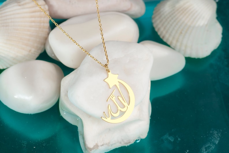 Peut inclure: Un collier dor&eacute; compos&eacute; d'une cha&icirc;ne fine et d'un pendentif en forme de croissant de lune surmont&eacute; d'une &eacute;toile. Le pendentif pr&eacute;sente la calligraphie arabe du mot Allah int&eacute;gr&eacute;e au design du croissant de lune, formant un bijou coh&eacute;rent.