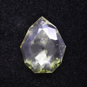 May include: A faceted, clear gemstone with a yellow-green hue. The gem is cut into a unique, geometric shape, with multiple facets that reflect light. The gemstone is set against a black background, highlighting its clarity and color.