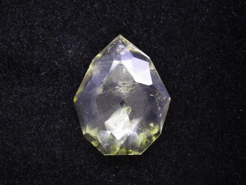 May include: A faceted, clear gemstone with a yellow-green hue. The gem is cut into a unique, geometric shape, with multiple facets that reflect light. The gemstone is set against a black background, highlighting its clarity and color.