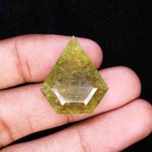 May include: A faceted, olive-green gemstone with a unique geometric shape. The gem is held in a hand against a black background. The stone has a translucent quality, with visible internal structures.