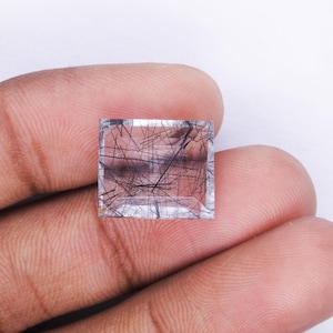 Pode incluir: Uma gema de quartzo transparente com inclusões pretas, cortada em forma quadrada. A gema é segurada na mão.