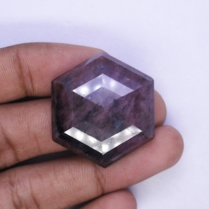May include: A hexagonal gemstone with a deep purple colour and faceted surfaces. The gem's geometric design reflects light, producing a sparkling effect. The stone is held in a hand, showing its size and shape.