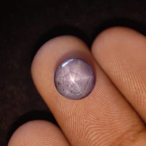 May include: A round, cabochon-cut star sapphire gemstone with a light purple hue. The stone displays a distinct six-rayed star effect across its polished, domed surface, with subtle natural inclusions visible near the edge of the gemstone.