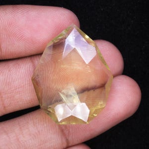 May include: A faceted, yellow-toned gemstone with a geometric shape. The gem is held between fingers against a dark background, showcasing its clarity and multiple facets. The gemstone's color ranges from pale yellow to a golden hue.