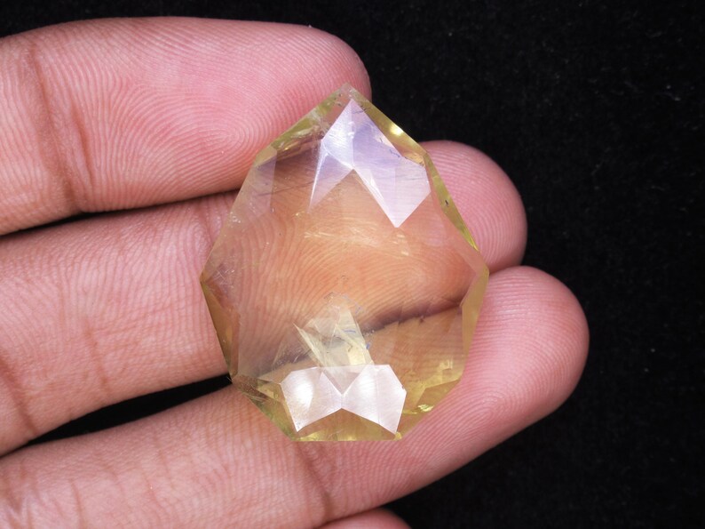 May include: A faceted, yellow-toned gemstone with a geometric shape. The gem is held between fingers against a dark background, showcasing its clarity and multiple facets. The gemstone's color ranges from pale yellow to a golden hue.