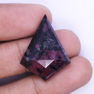 May include: A faceted gemstone with a kite shape, held in a hand. The gem features a deep purple and black color with a reflective surface. The gem is a unique shape and design.