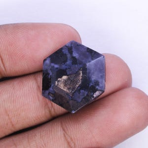 May include: A faceted, hexagonal gemstone with a deep blue and black color scheme, featuring a central area of gold-colored inclusions. The gem is held in a hand, showcasing its polished surfaces and intricate internal patterns. The gem's shape is geometric.