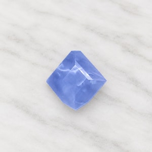 May include: A faceted periwinkle blue gemstone with a kite-like shape. The stone features a smooth, polished surface with multiple angular cuts that create light reflections across its translucent, cool-toned blue body.