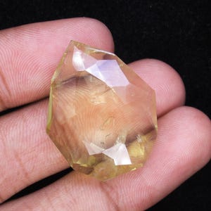 May include: A faceted, light yellow gemstone with a geometric shape. The gem is held in a hand against a dark background, showcasing its clarity and color. The cut of the stone creates multiple reflective surfaces.