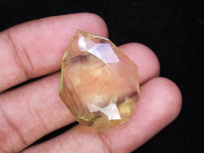 May include: A faceted, light yellow gemstone with a geometric shape. The gem is held in a hand against a dark background, showcasing its clarity and color. The cut of the stone creates multiple reflective surfaces.