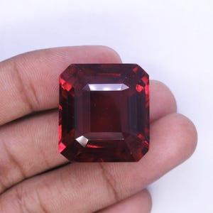 Op de afbeelding: Een grote, rechthoekige, gefacetteerde edelsteen met een dieprode kleur. De edelsteen wordt in een hand gehouden, waardoor de gepolijste oppervlakken en interne reflecties worden benadrukt. De slijpvorm is een smaragd, met een symmetrische rangschikking van facetten.