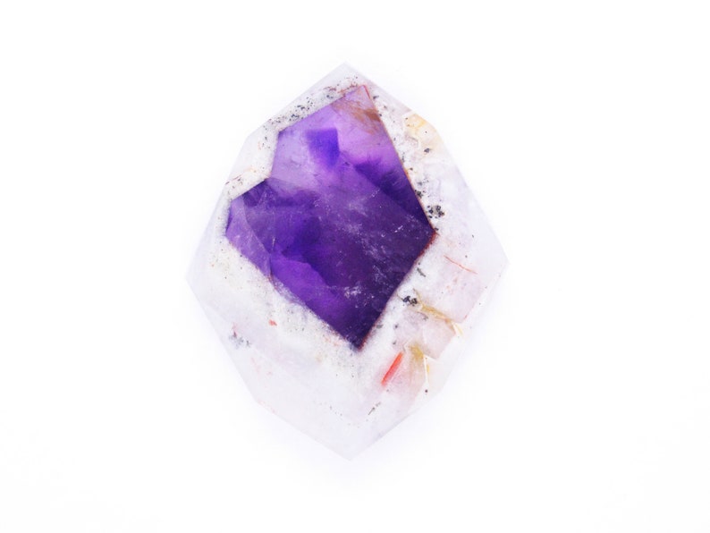 Pu&ograve; includere: Un cristallo di ametista viola con un esterno chiaro, bianco e rosa. Il cristallo ha la forma di una punta e una texture ruvida e naturale.