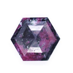 Op de afbeelding: Een gefacetteerde edelsteen met een zeshoekige vorm. De edelsteen heeft een mix van kleuren, waaronder diep roze, zwart en wit. De steen staat tegen een witte achtergrond, wat de unieke kleurvariaties en geometrische vorm benadrukt.