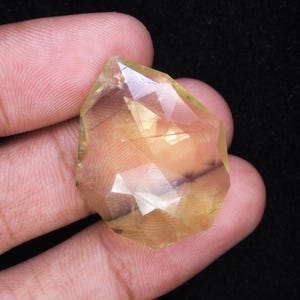 May include: A faceted, light yellow gemstone with a complex, geometric cut. The gem is held in a hand against a dark background, showcasing its clarity and color. The stone's facets reflect light, creating a sparkling effect.