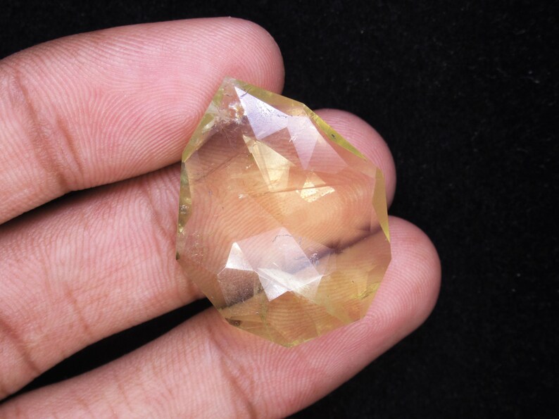 May include: A faceted, light yellow gemstone with a complex, geometric cut. The gem is held in a hand against a dark background, showcasing its clarity and color. The stone's facets reflect light, creating a sparkling effect.