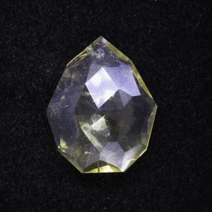 May include: A faceted, teardrop-shaped gemstone with a pale yellow hue. The crystal is transparent, with multiple geometric facets that reflect light. The gem is set against a black background, highlighting its clarity and shape.