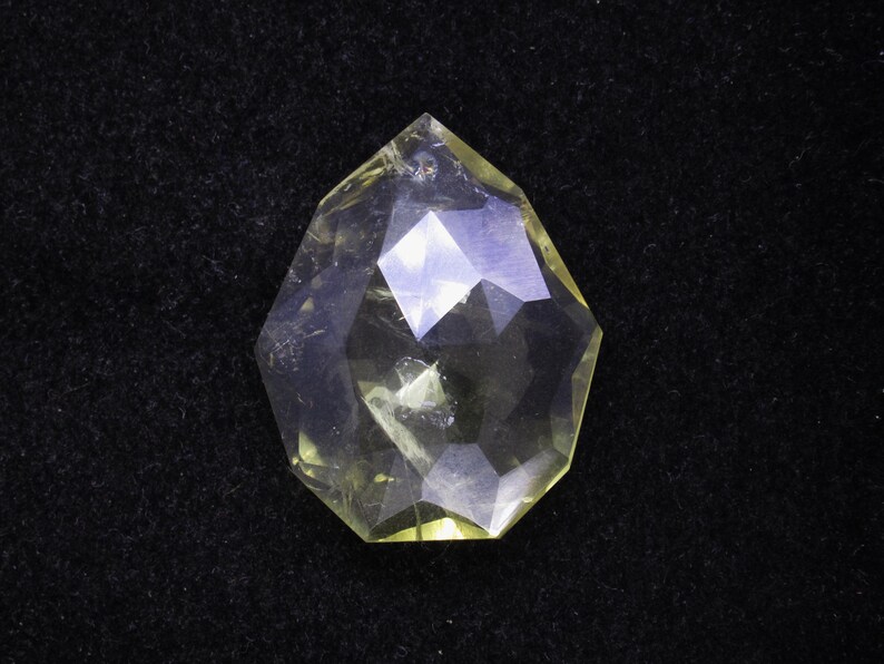 May include: A faceted, teardrop-shaped gemstone with a pale yellow hue. The crystal is transparent, with multiple geometric facets that reflect light. The gem is set against a black background, highlighting its clarity and shape.