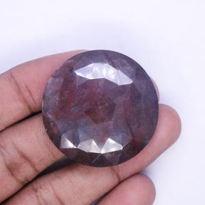 May include: A large, faceted, round gemstone with a deep burgundy color. The gem has multiple facets that catch the light, creating a sparkling effect. The stone is held in a hand against a white background.