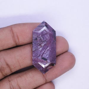 May include: A faceted, elongated purple gemstone with internal veining and a polished finish. The crystal is held in a hand, showcasing its geometric shape and color variations. The gemstone's facets catch the light, highlighting its depth and clarity.