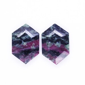 Puede incluir: Dos cabujones de piedras preciosas facetadas con forma hexagonal. Las piedras muestran una mezcla de colores, incluyendo rojo intenso, negro y verde. Las piedras preciosas están sobre un fondo blanco.
