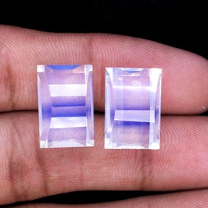 Puede incluir: Dos piedras preciosas rectangulares transparentes con un brillo iridiscente azul claro. Las piedras preciosas son facetadas y tienen una forma rectangular.