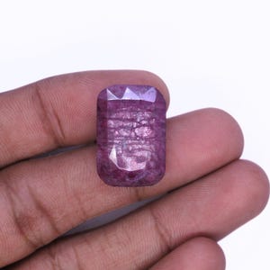 Peut inclure: Une pierre précieuse taillée, de forme rectangulaire, de couleur rouge violacé. La gemme est tenue dans une main sur fond blanc. La surface de la pierre présente des textures internes et des facettes qui réfléchissent la lumière.