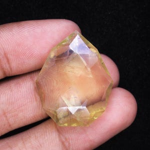 May include: A faceted, yellow-toned gemstone with a complex, geometric cut. The gem is held in a hand against a dark background, showcasing its clarity and light refraction. The gem's shape is a modified hexagon.