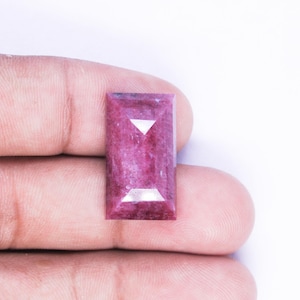 Könnte beinhalten: Ein rechteckiger facettierter Edelstein in einem tiefen Magenta- und Rosaton. Der Stein hat eine flache Oberseite mit abgeschrägten Kanten, die ein geometrisches Muster bilden und das Licht auf seiner polierten, halbtransparenten Kristallstruktur reflektieren.