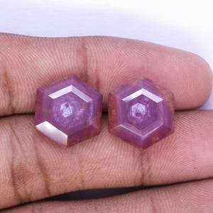 May include: Two faceted, hexagonal gemstones with a deep pink hue. Each gem features a central, smaller hexagon within the larger shape. The gems are displayed against a neutral background, highlighting their color and cut.