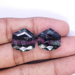 Può includere: Due gemme sfaccettate a forma esagonale. Le gemme hanno un colore di base scuro con inclusioni rosse e verdi. Le sfaccettature riflettono la luce, creando un effetto scintillante. Le gemme sono esposte su uno sfondo neutro.