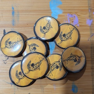 May include: A collection of round, wooden-toned buttons with a black border. Each button features a black illustration of a winged figure. The buttons are arranged on a wooden surface with paint splatters.