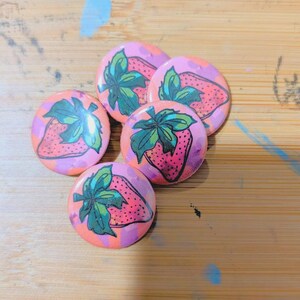 May include: A collection of five round buttons featuring a hand-drawn strawberry design. The strawberries are pink with green leaves and are set against a background of orange and purple. The buttons are arranged on a wooden surface.