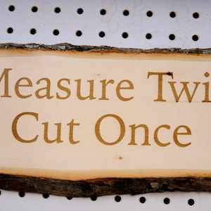 May include: A wooden sign with a rustic bark edge, featuring the text "Measure Twice Cut Once" in a light brown color.