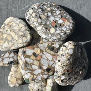 May include: A collection of smooth, rounded stones featuring a terrazzo-style mosaic pattern. Each stone displays a dense mix of small, angular fragments in shades of white, grey, black, tan, and occasional orange embedded within a solid base.