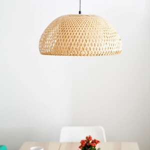 May include: A woven bamboo pendant light hangs above a table with a small potted plant. The light fixture has a dome shape with a lattice design. The plant has red flowers and is in a woven basket.