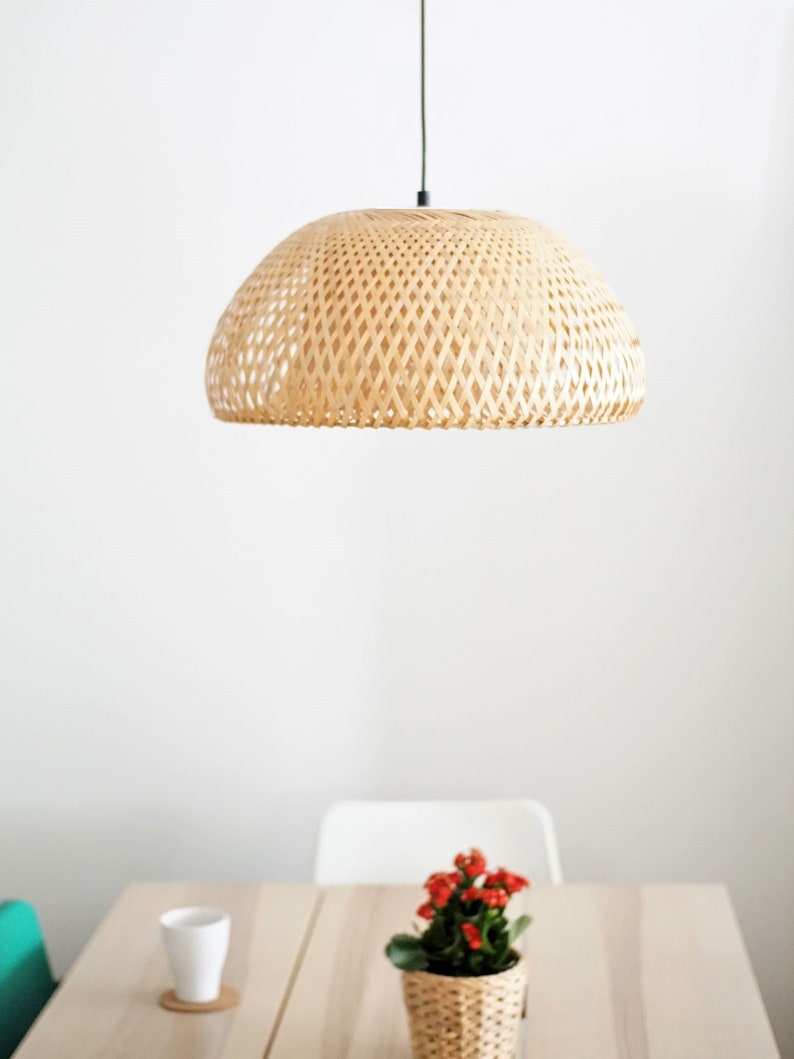 May include: A woven bamboo pendant light hangs above a table with a small potted plant. The light fixture has a dome shape with a lattice design. The plant has red flowers and is in a woven basket.