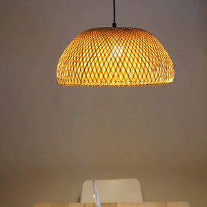 May include: A woven bamboo pendant light fixture hangs above a wooden table. The light is dome-shaped and emits a warm glow. A laptop, a white mug, and a coaster are on the table. The background is a neutral color.