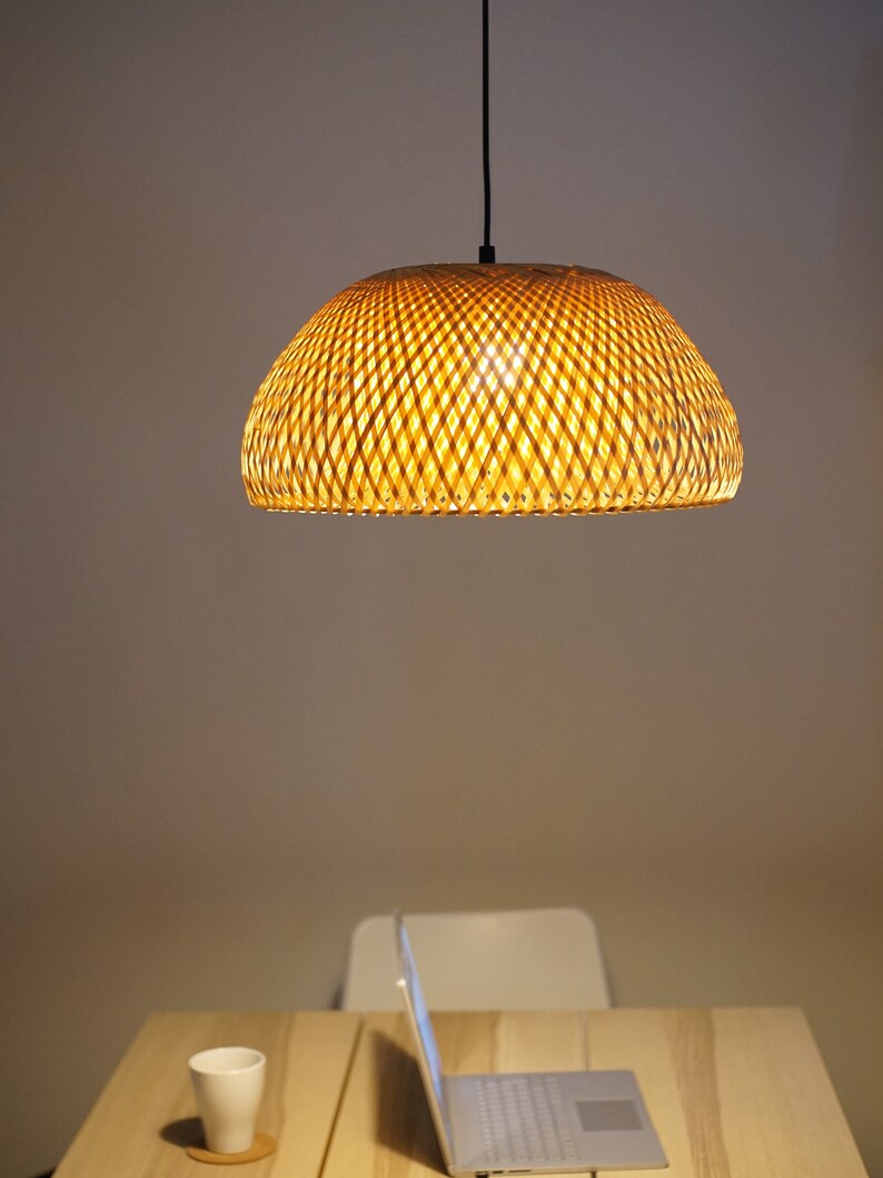 May include: A woven bamboo pendant light fixture hangs above a wooden table. The light is dome-shaped and emits a warm glow. A laptop, a white mug, and a coaster are on the table. The background is a neutral color.