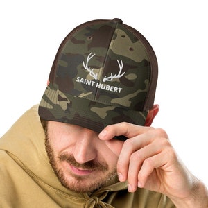 May include: A camouflage trucker hat with a brown mesh back. The hat has a white embroidered deer antler logo with the text "SAINT HUBERT" underneath.
