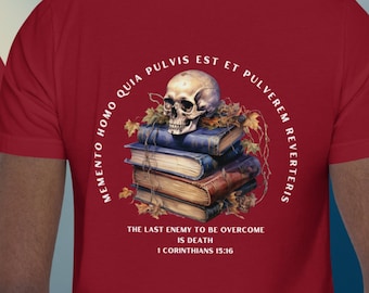 May include: A maroon t-shirt with the words "MEMENTO MORI" printed in white on the front. The back features a skull atop a stack of books, with Latin text and the phrase "THE LAST ENEMY TO BE OVERCOME IS DEATH" and the Bible verse 1 Corinthians 15:16.