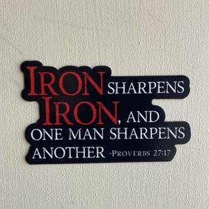 May include: A black sticker with the text "IRON SHARPENS IRON, AND ONE MAN SHARPENS ANOTHER -PROVERBS 27:17" in red and white. The sticker has a unique shape and is likely for decorative purposes.