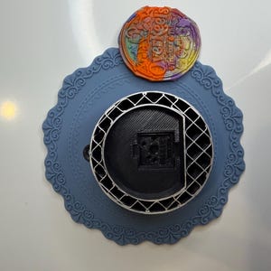 Può includere: Un sottobicchiere circolare in silicone azzurro con un bordo smerlato decorativo. Al centro si trova un oggetto circolare nero stampato in 3D con una struttura a griglia. Sopra è posizionato un piccolo gettone rotondo multicolore con il testo OverKeels.