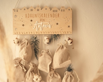 May include: A wooden wall-hanging advent calendar with the text ADVENTSKALENDER VON Antonia, featuring a skyline cutout, star details, and numbers 1 through 24. Nine beige fabric drawstring pouches hang from the board, decorated with cotton sprigs.