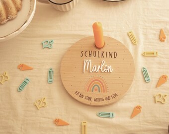 May include: A round wooden plaque featuring the text "SCHULKIND Marlon" and "ICH BIN STARK, WICHTIG UND KLUG" with a rainbow graphic and a central hole for a candle. Scattered around are small wooden shapes of pencils, carrots, and the number 123.