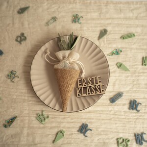 May include: A brown burlap cone filled with green foliage and tied with a white ribbon. The cone is sitting on a beige plate with a wooden sign that reads "Erste Klasse". The plate is surrounded by small green and blue decorations.