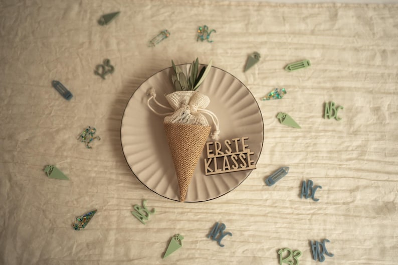 May include: A brown burlap cone filled with green foliage and tied with a white ribbon. The cone is sitting on a beige plate with a wooden sign that reads "Erste Klasse". The plate is surrounded by small green and blue decorations.