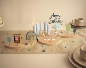May include: A wooden birthday ring centrepiece featuring a central circular base with the name Hannes, the number 10, and four light blue candles. It includes decorative wooden inserts of a rainbow, a photo frame, balloons, and a Happy Birthday sign.