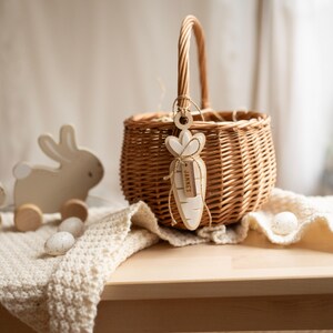 May include: A wicker basket with a wooden carrot tag that says "Jamie's" hanging from the handle. The basket is sitting on a white crocheted blanket with two wooden bunny toys and white eggs in front of it.