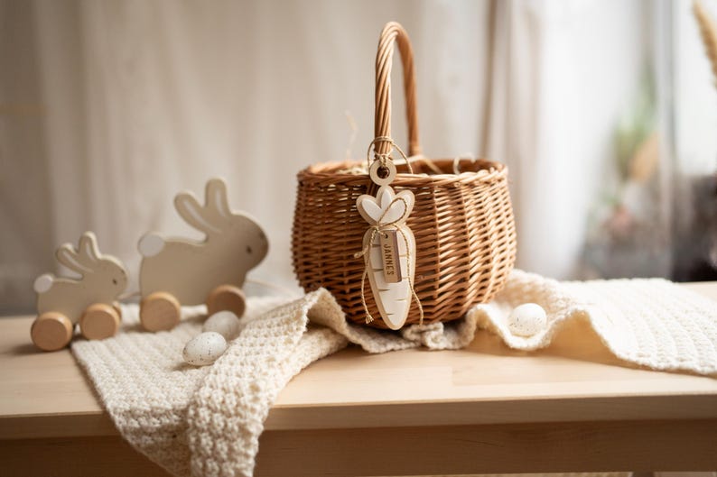May include: A wicker basket with a wooden carrot tag that says "Jamie's" hanging from the handle. The basket is sitting on a white crocheted blanket with two wooden bunny toys and white eggs in front of it.